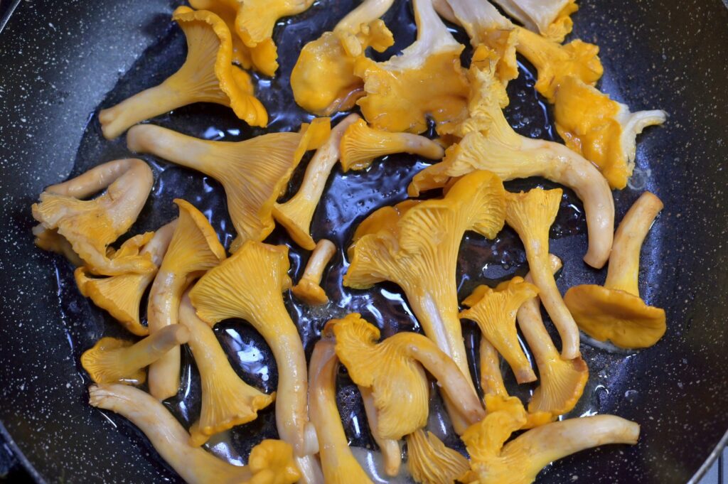 Freshly cleaned chanterelle mushrooms sizzling in a pan with oil, showcasing their delicate ridges and rich yellow-orange hue. The mushrooms are lightly cooking, releasing moisture and beginning to brown slightly.
