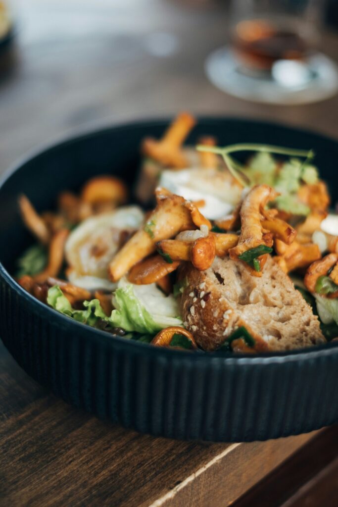 A rustic salad featuring sautéed chanterelle mushrooms, crusty bread chunks, and leafy greens, served in a black ribbed bowl. The focus is on the golden mushrooms nestled among the fresh and hearty ingredients.
