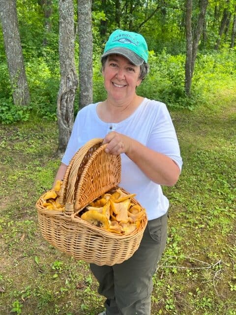 Nancy Etzwiler wearing a turquoise "Grandma's" baseball cap and white shirt stands in a forest clearing, holding a woven basket full of golden chanterelle mushrooms. The scene is lush and green, with tall trees and dappled sunlight in the background.