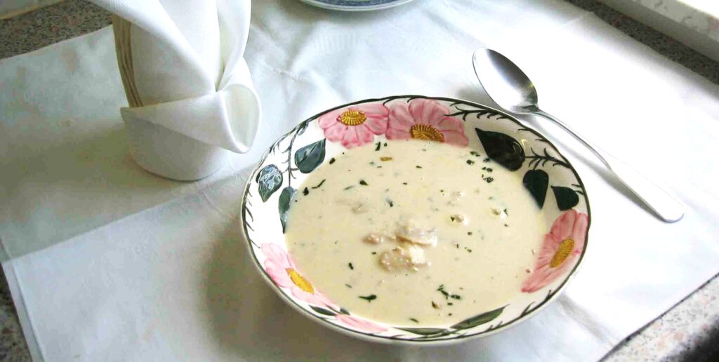 A bowl of creamy mushroom soup garnished with herbs, served on a floral-patterned dish atop a white placemat with a spoon and folded napkin beside it. The soup appears homemade with chunks of mushrooms visible in the thick, white broth.