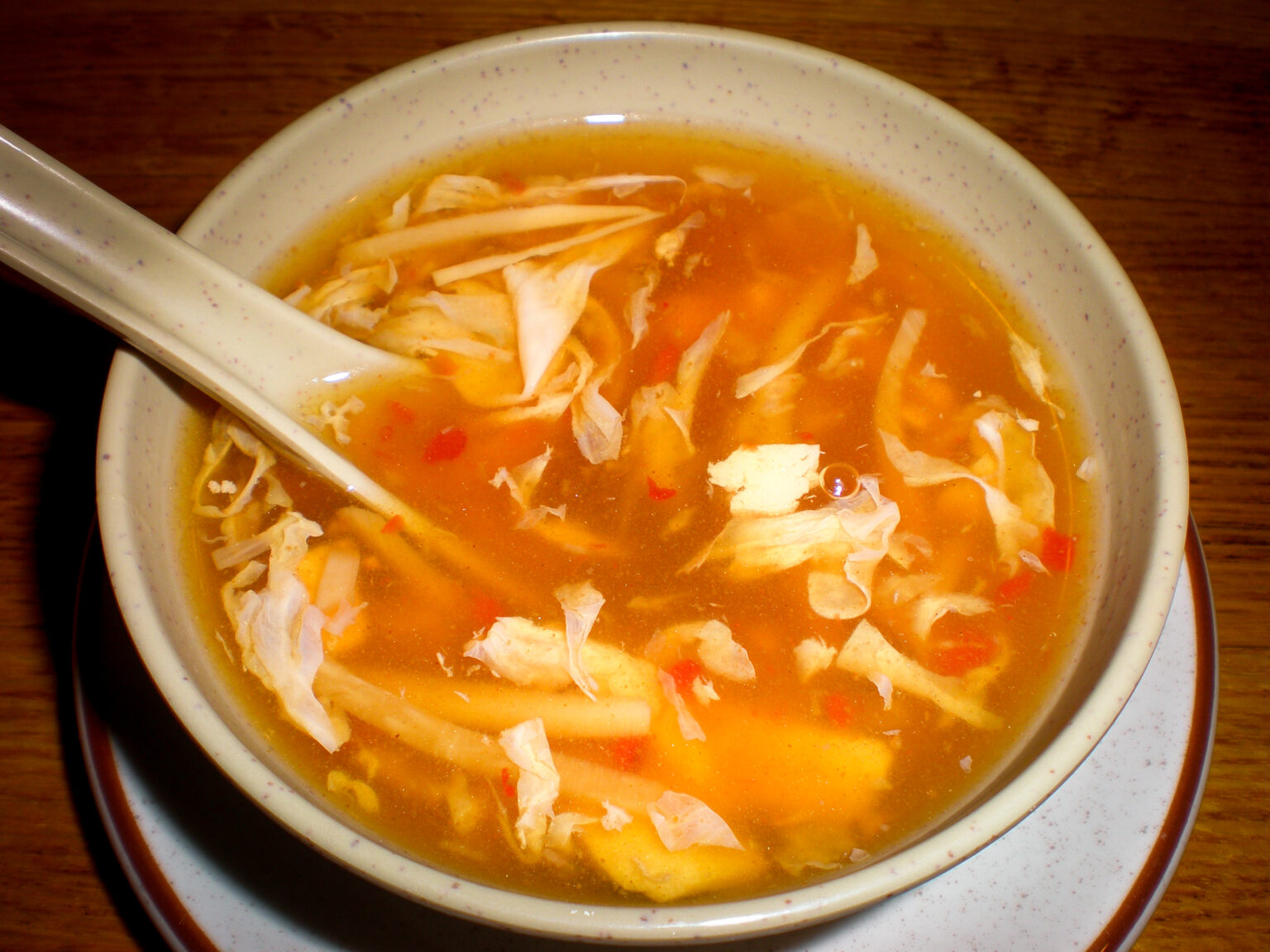A bowl of hot and sour soup featuring strips of tofu, bamboo shoots, and red peppers in a tangy orange broth. A ceramic soup spoon rests in the bowl, which is placed on a matching saucer atop a wooden table.