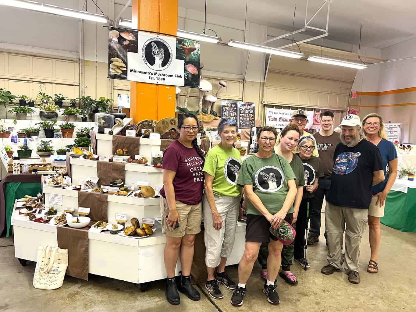 A group of Minnesota Mycological Society volunteers pose in front of their mushroom display booth at the Minnesota State Fair. The display features labeled mushroom specimens arranged on white tiered platforms with brown cloth runners. Behind them hangs a banner reading "Minnesota’s Mushroom Club Est. 1899," surrounded by plants and educational materials.