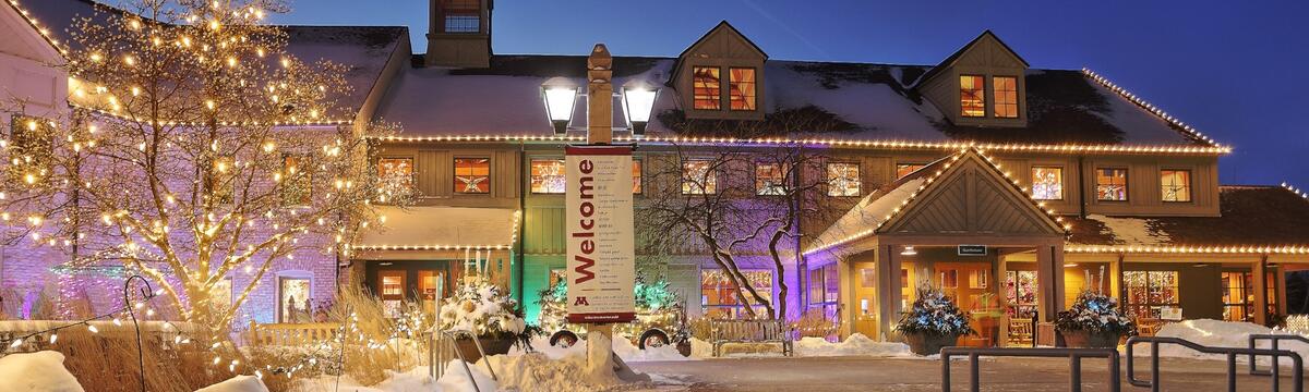 Exterior of the Minnesota Landscape Arboretum’s Oswald Visitor Center at dusk in winter, decorated with glowing string lights and snow-covered trees. A large ‘Welcome’ banner hangs near the entrance, inviting visitors to the Winter Lights event.