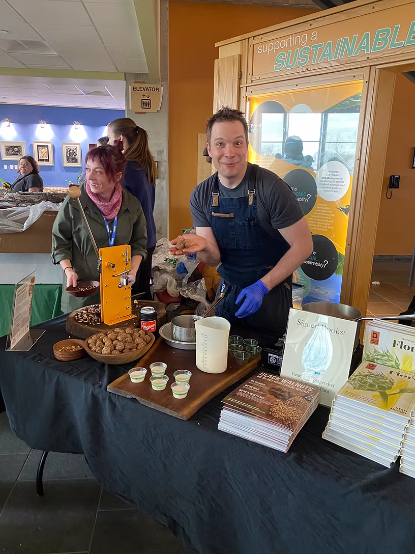 Chef and forager Alan Bergo smiles at the camera while preparing a black walnut demonstration at an event booth, wearing a denim apron and blue gloves. A wooden board holds walnuts, small tasting cups, and a measuring cup, with signed copies of his books displayed at the front of the table. A volunteer with a pink scarf works alongside him in the background.