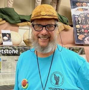 Ron Spinosa, wearing a brown hat and a blue volunteer shirt, smiles in front of the Minnesota Mycological Society display at the 2022 State Fair, with oversized model mushrooms and educational posters behind him.