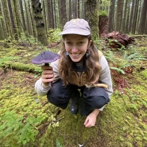 Elora Adamson squatting in a moss ground covered forest smiling while holding a mushroom in one hand.